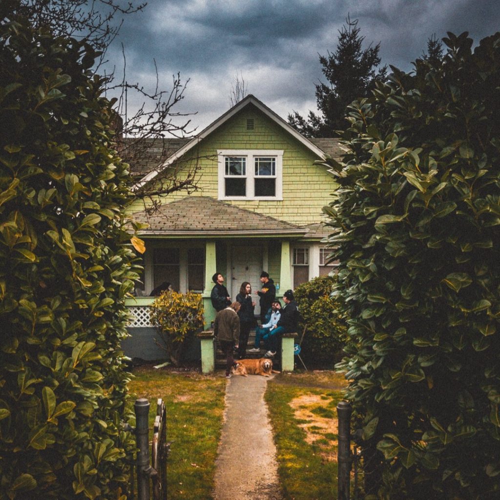 Portal de casa al fondo de un jardín con amigos conviviendo y un perro echado al frente.