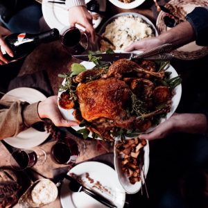 Una mesa donde hay platos y copas en la parte del fondo, y en primer plano unas manos se pasan un platón con un pollo rostizado.