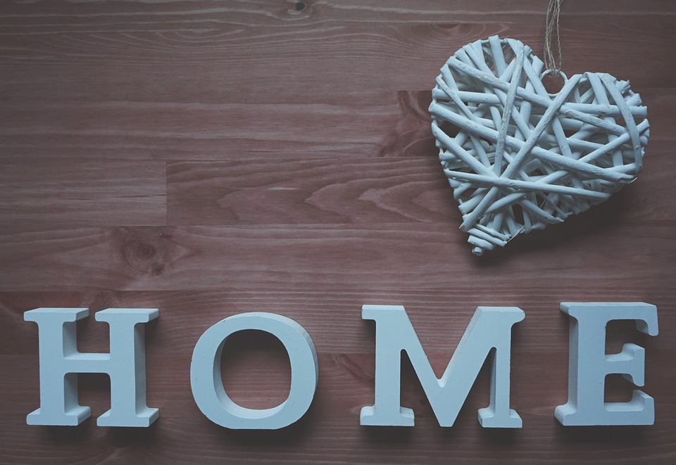 Sobre fondo de madera la palabra "HOME" hogar en ingles y un corazón trenzado ambos en blanco.