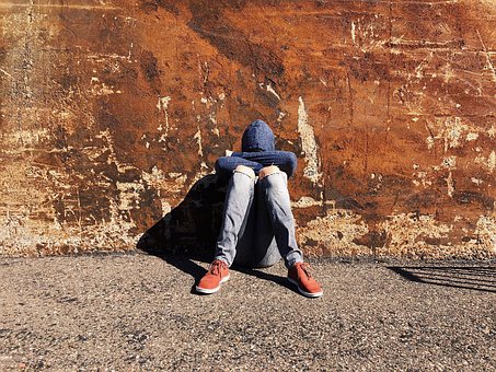 joven sentado en la calle, apoyado contra la pared con la cabeza hundida entre los brazos apoyados cobre las piernas.