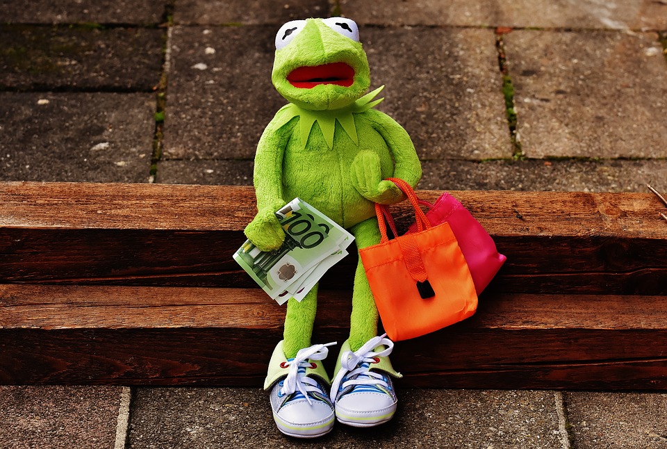 La rana Rene de los Muppets sentada, con un billete de 100 euros y dos bolsas de compras y expresión de desaliento.